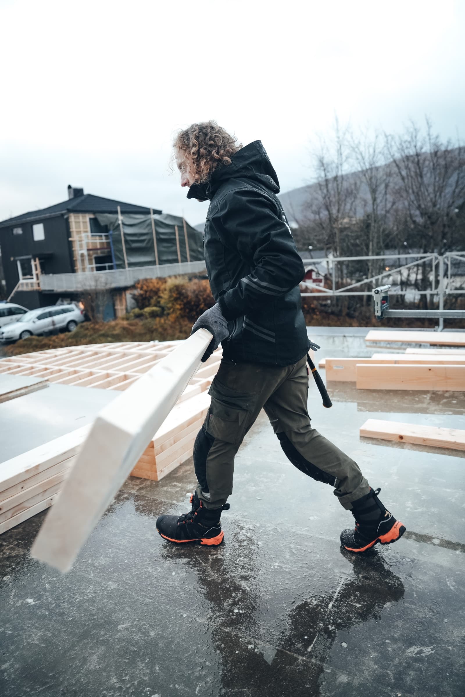 Tømrer bærer materialer over en nystøpt plate på en byggeplass med boliger i bakgrunnen